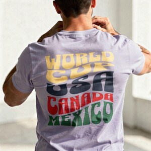 May include: A light purple t-shirt with the words "WORLD CUP USA CANADA MEXICO" in a retro font. The text is in yellow, navy, red, and green. The shirt is being worn by a person.