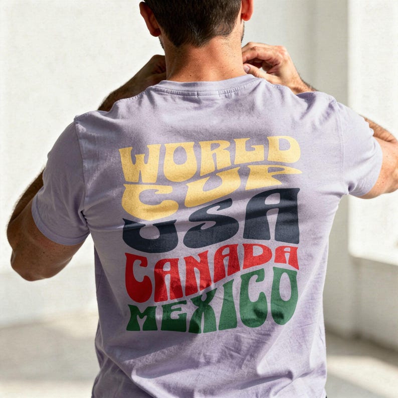 May include: A light purple t-shirt with the words "WORLD CUP USA CANADA MEXICO" in a retro font. The text is in yellow, navy, red, and green. The shirt is being worn by a person.