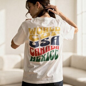 May include: White t-shirt with a colorful graphic on the back. The graphic reads "WORLD CUP USA CANADA MEXICO" in yellow, blue, red, and green, with a retro font. The shirt is short-sleeved and appears to be made of cotton.