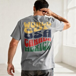 May include: A gray t-shirt with the words "WORLD CUP USA CANADA MEXICO" in a retro font across the back. The text is in yellow, navy blue, red, and green. The shirt is short-sleeved.