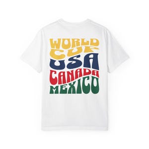 May include: White t-shirt with a colorful graphic on the back. The graphic reads "WORLD CUP USA CANADA MEXICO" in a retro font, with each word in a different color: yellow, blue, red, and green.