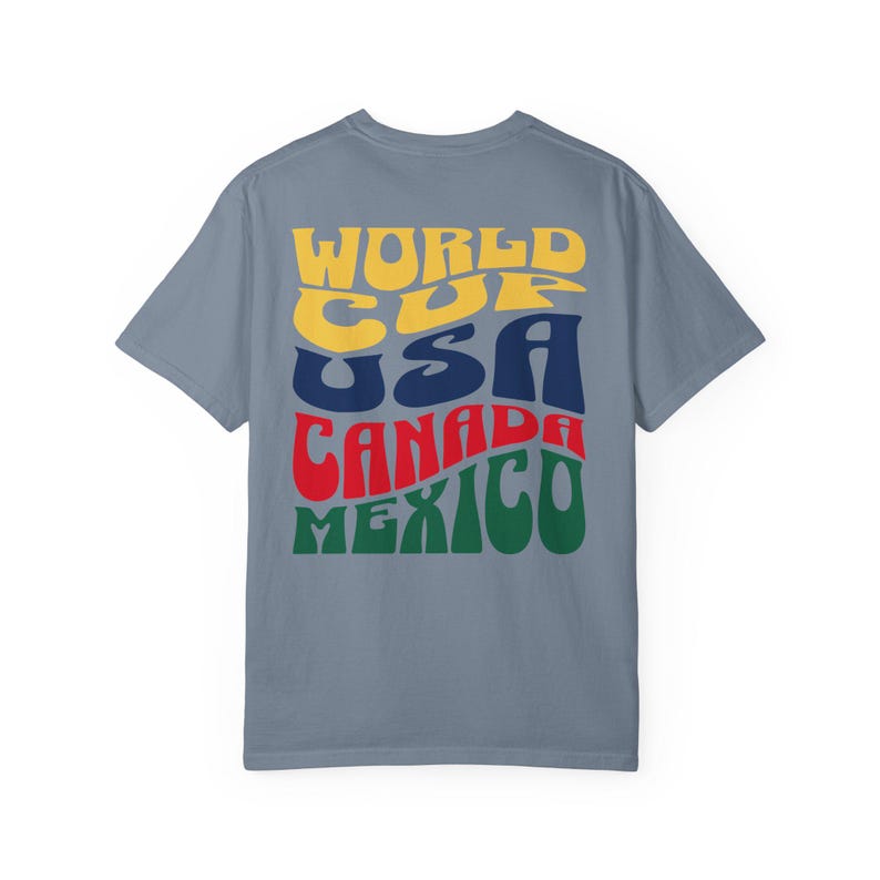 May include: A blue t-shirt with the words "WORLD CUP USA CANADA MEXICO" in a retro font. The text is in yellow, blue, red, and green. The shirt is a solid color and has short sleeves.