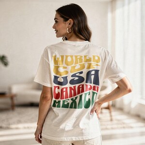May include: Cream-colored t-shirt with the words "WORLD CUP USA CANADA MEXICO" printed on the back in yellow, navy, red, and green. The shirt has short sleeves and a relaxed fit. The person is wearing gold hoop earrings.