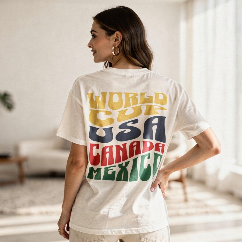 May include: Cream-colored t-shirt with the words "WORLD CUP USA CANADA MEXICO" printed on the back in yellow, navy, red, and green. The shirt has short sleeves and a relaxed fit. The person is wearing gold hoop earrings.