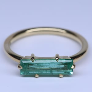 May include: A gold ring featuring a horizontal, rectangular, baguette-cut green gemstone set in a four-prong setting. The band is smooth and polished, showcasing a simple, clean design that highlights the elongated shape of the vibrant green stone.