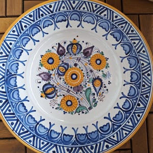 Puede incluir: Plato de cerámica decorativo con diseño floral. El plato presenta un centro blanco con flores amarillas y azules, rodeado por un borde azul y blanco y un borde amarillo. El plato mide aproximadamente 30 cm de diámetro.