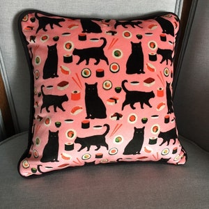 Op de afbeelding: Vierkant kussen met een roze achtergrond met een herhaald patroon van zwarte kat silhouetten en sushi rollen. Het kussen heeft een donkerbruine rand en ligt op een grijze stoel.