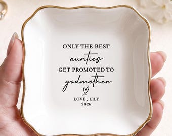 May include: A square white ceramic trinket dish with a scalloped edge and a gold-painted rim. The center features black text that reads: ONLY THE BEST aunties GET PROMOTED TO godmother, followed by a small heart symbol, LOVE, LILY, and 2026.