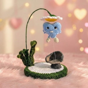 Puede incluir: Una escultura de ganchillo caprichosa con un personaje de corazón azul suspendido de una flor. La base es verde y blanca con una seta. El fondo es rosa suave con bokeh en forma de corazón. Un objeto decorativo único.