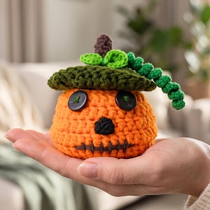 May include: A small, round, orange crocheted pumpkin figure with a dark green crocheted hat, a brown stem, and a curly green vine. The pumpkin features two black button eyes, a black stitched triangular nose, and a horizontal black stitched mouth.