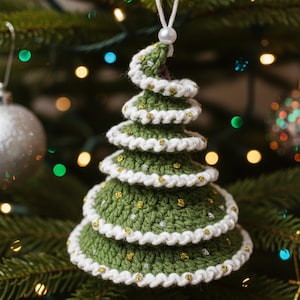 Peut inclure: Ornement de Noël fait à la main en crochet vert, avec des bordures blanches et des accents dorés. L'arbre à plusieurs niveaux a un sommet en spirale et une ficelle blanche pour l'accrocher. L'ornement est sur un sapin de Noël.