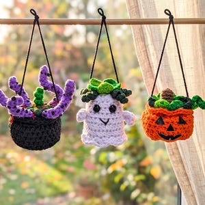 May include: Three hanging crochet decorations suspended from a rod. The left is a black pot with purple tentacles. The center is a white ghost with green foliage on top. The right is an orange jack-o'-lantern with green leaves and a vine.