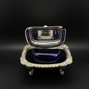 以下が含まれることがあります： ヒンジ付きの蓋が付いた、銀色の装飾的な皿。深い青色の内側が特徴です。正方形の皿には装飾的な縁があり、小さく湾曲した足の上に置かれています。蓋は開いており、青色の内側が見えます。