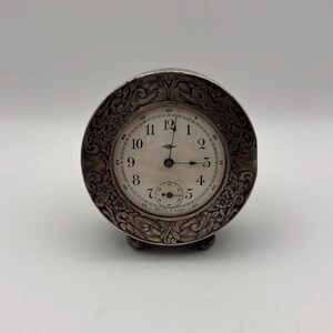 May include: A silver-toned antique clock with an ornate floral design around the face. The clock face is white with black numerals and hands. The clock has a small seconds dial and rests on small feet.