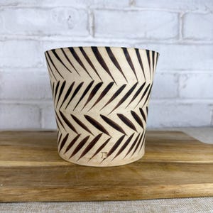 May include: A cylindrical ceramic planter featuring a cream-colored base with a repeating dark brown chevron or leaf-like pattern arranged in horizontal bands. The vessel has a smooth finish and a small square maker's mark near the bottom edge.