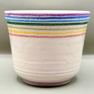 Puede incluir: Una maceta de cerámica blanca con una serie de rayas horizontales texturizadas en morado, azul, verde, amarillo y rojo cerca del borde superior. La base de la maceta también muestra una fina raya morada y una textura ligeramente irregular.