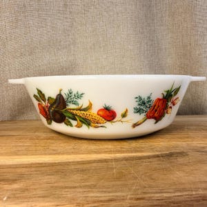 May include: A white milk glass mixing bowl with two side handles, featuring a colourful wrap-around illustration of various vegetables including a dark pear, yellow corn, a red tomato, and green herbs printed along the exterior surface.