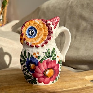 May include: A ceramic owl-shaped jug featuring a hand-painted design with a large orange eye, a white body, and vibrant floral patterns in shades of pink, blue, and green. The jug includes a curved handle and a sculpted owl head top.