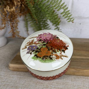 May include: A round, cream-coloured porcelain trinket box with a lid featuring a floral design of pink, red, orange, and purple chrysanthemums. The base of the box is decorated with a repeating reddish-brown scalloped pattern and gold trim.