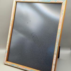 Könnte beinhalten: Ein rechteckiger Bilderrahmen mit einem goldenen Innenrand und einem dekorativen Außenrand mit einem pastellfarbenen Blumenmuster in Rosa, Blau und Gold. Der Rahmen hat eine schwarze Rückseite und wird von einem schwarzen Ständer gehalten.