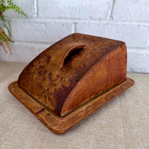 May include: A rectangular brown ceramic butter dish with a domed lid featuring an embossed floral pattern and a central arched handle. The base has a matching embossed border, and the entire piece has a rustic, textured finish in earthy tones.