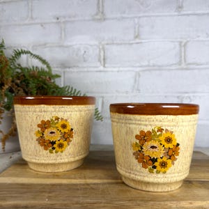 May include: Two ceramic planters with a speckled cream finish and a brown glazed rim. Each planter features a central floral decal displaying clusters of yellow, orange, and brown flowers. The planters have a cylindrical shape with a slightly tapered base.