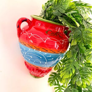 May include: A wall-mounted ceramic vase featuring a vibrant red upper section with white painted swirls, a central blue band with white floral-like patterns, and a red base. The vase has two side handles and contains a green artificial plant.