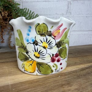 May include: A white ceramic planter with a ruffled top edge, hand-painted with a floral motif featuring white, yellow, and pink blossoms, green leaves, and small blue and red accents. The design includes a signature near the base of the pot.