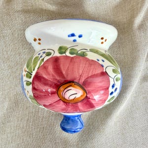 May include: A ceramic wall pocket planter featuring a hand-painted design of a large red flower with an orange centre. The piece is decorated with green leaves, blue dots, and brown accents on a white background, finished with a blue base knob.