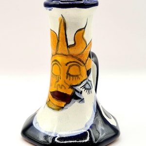 May include: A ceramic candle holder with a white base and a dark blue rim and bottom. The front features a hand-painted yellow sun motif with closed eyes and a dark red mouth. The piece has a flared shape and a small handle on the right side.