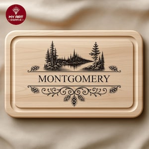 Op de afbeelding: Lichtgekleurde houten snijplank met een gegraveerd ontwerp. Het ontwerp bevat een landschap met bomen, een rivier en de naam "MONTGOMERY" in een decoratief lettertype. De plank heeft een verzonken rand. Afmetingen in centimeters.