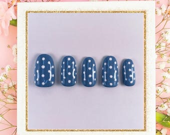 May include: Five blue press-on nails with white polka dots arranged in a row. The nails are oval-shaped and set against a white background with a gold border, surrounded by pink and white flowers.