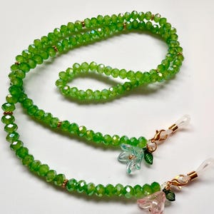May include: A green beaded eyeglass chain featuring faceted glass beads, small gold-toned spacers, and two clear silicone loops at the ends. The chain is accented with two small flower-shaped charms in light blue and pink with green leaves.