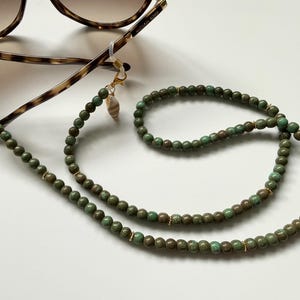 May include: A beaded eyeglass chain featuring round, mottled green and brown beads interspersed with small gold-tone spacers. The chain attaches to tortoise-shell patterned sunglasses with clear silicone loops and gold-tone lobster clasps, accented by small seashell charms.
