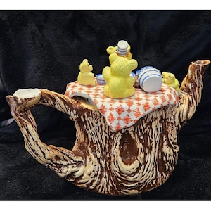 May include: A whimsical teapot shaped like a tree stump, featuring a handle and spout. The lid is adorned with yellow bear figurines and miniature tea party accessories. The teapot has a brown and beige wood-grain design, with a red and white checkered tablecloth.