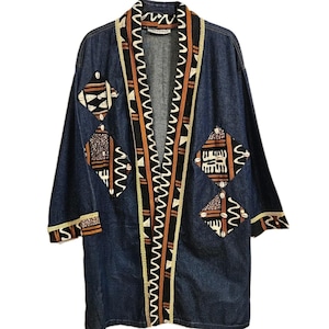 May include: A dark blue denim jacket with an open front and long sleeves. The jacket features decorative patches with geometric patterns in brown, black, and white. The collar and cuffs are trimmed with patterned fabric. The jacket has a relaxed fit.