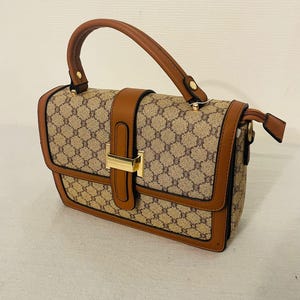 May include: A rectangular handbag with a brown handle and trim. The bag features a repeating pattern in tan and brown, with a gold-colored clasp. The bag has a structured design and a top handle.