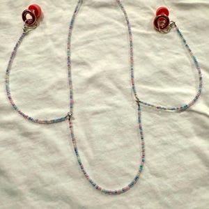 May include: A beaded eyeglass chain with two red earbud holders. The chain is made of small, colourful beads in shades of pink, blue, and purple. The chain is designed to hold eyeglasses or sunglasses.