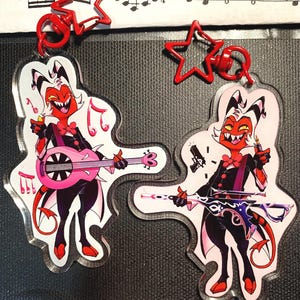 May include: Two clear acrylic keychains featuring a cartoon character with red skin, black clothing, and a white hairstyle. One keychain has a pink guitar, the other a gun. Both have red star-shaped clasps. Musical notes are visible.