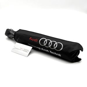 Puede incluir: Un paraguas negro enrollado con el logotipo de Audi en plata y la palabra "Audi" en rojo. También se ve el texto "Vorsprung durch Technik". Una etiqueta blanca con el logotipo de Audi está adjunta.