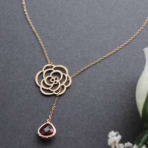 Könnte beinhalten: Eine goldfarbene Lariat-Halskette mit einer feinen Kette, einem zentralen Anhänger in Form einer durchbrochenen Blume und einem einzelnen tropfenförmigen, facettierten dunkelroten Edelsteinanhänger am unteren Ende.