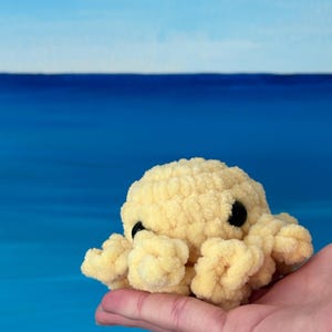 Könnte beinhalten: Ein kleines, gelbes Plüsch-Oktopus mit schwarzen Augen. Der Oktopus besteht aus einem weichen, strukturierten Material und hat acht gekräuselte Tentakel. Der Hintergrund ist ein Blauton, der an das Meer und den Himmel erinnert.