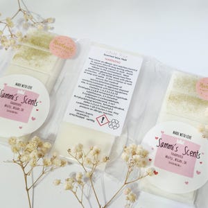 Op de afbeelding: Drie pakketten geurwasmelts, elk met het label "Sammi's Scents". De witte wasmelts hebben een lichte groene stoflaag. De pakketten zijn verzegeld in helder plastic en versierd met roze hartstickers en gedroogde bloemen.