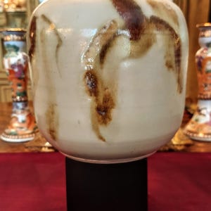 Könnte beinhalten: Eine zylindrische Keramikvase mit einer glatten, cremeweißen Grundglasur und unregelmäßigen, dunkelbraunen vertikalen Tropfenmustern. Die Vase hat einen abgerundeten oberen Rand und steht auf einem soliden, quadratischen schwarzen Sockel.