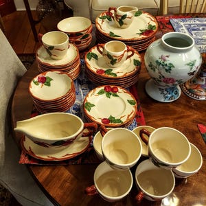 May include: A collection of vintage ceramic tableware with a red apple design on a cream background. The set includes cups, saucers, plates, bowls, and a creamer. The rims and handles are accented with a red trim, creating a classic aesthetic.