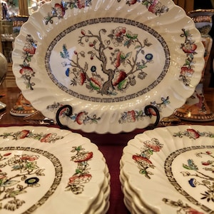 May include: An oval serving platter and several matching plates. The white ceramic pieces feature a floral and tree design in red, blue, and green, with a brown border. The edges have a scalloped design.
