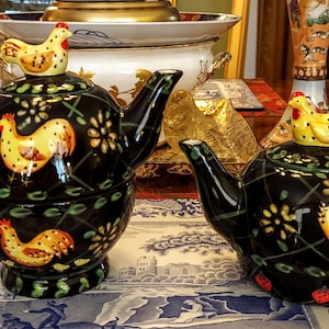 May include: Two black ceramic teapots with chicken designs. One teapot has a cup attached. The teapots have yellow chicken figures on the lids and chicken illustrations on the body. The teapots are decorated with floral and vine patterns.