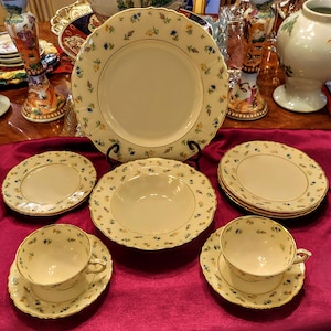 Puede incluir: Un juego de vajilla de porcelana crema con motivos florales y ribete dorado. El juego incluye platos llanos, platos de ensalada, un cuenco para sopa y tazas de té con platillos. El estampado floral presenta pequeñas flores azules y amarillas. Sobre un mantel burdeos.