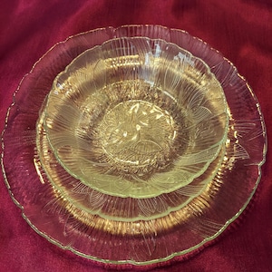 May include: Three clear glass plates of varying sizes are stacked on top of each other. The plates have a textured, floral design and scalloped edges. The plates are arranged on a deep red surface.