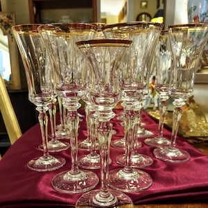 May include: A set of clear crystal champagne flutes with gold rims. The glasses have a tall, slender design with a decorative stem and a wide base. They are arranged on a burgundy surface, reflecting light.